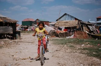 Cheerful girl on a bicycle in Cambodia in front of tin shacks.