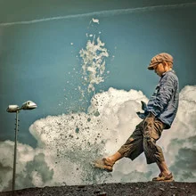 Fotokunst von Ambra A: Junge mit Schiebermütze spielt in einer Pfütze unter Wolken.