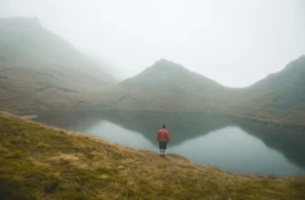 Photo art: Man by a tranquil lake, surrounded by mountains and fog in Scotland.