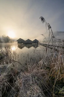 Three cabins at Lake Kochel in morning light with calm reflections on the water.