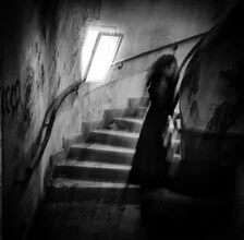 Black and white photo of a girl figure at the bottom of stairs in soft light.