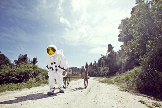 Astronaut pulls an empty wooden cart on a street under a cloudy sky.